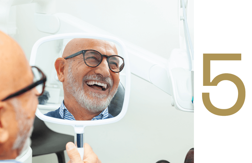 Smiling elderly man with glasses looking at his reflection in a dental mirror, showcasing satisfaction after dental implant procedure at Advanced Implant & Cosmetic Dentistry.