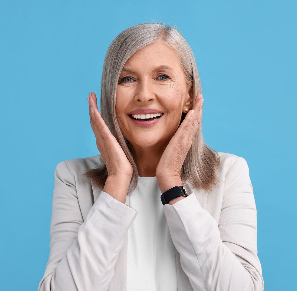 Smiling older woman with gray hair and hands on her cheeks, expressing joy, symbolizing successful dental implant results at Advanced Implant & Cosmetic Dentistry.