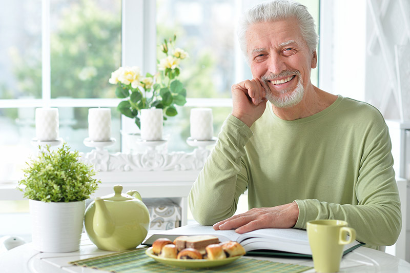 Smiling elderly man sitting at a table with a book, enjoying a cup of tea and pastries, in a bright, cozy environment, reflecting a sense of comfort and well-being.