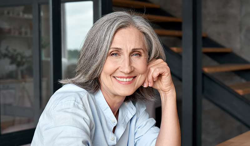 Smiling older woman with gray hair in a casual blue shirt, showcasing confidence and warmth, relevant to discussions on dental implants and patient care.