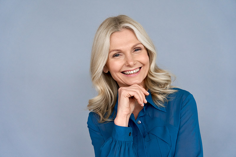Smiling middle-aged woman with long blonde hair, wearing a blue blouse, showcasing confidence and happiness, representing successful dental restoration in the context of implant-supported bridges.
