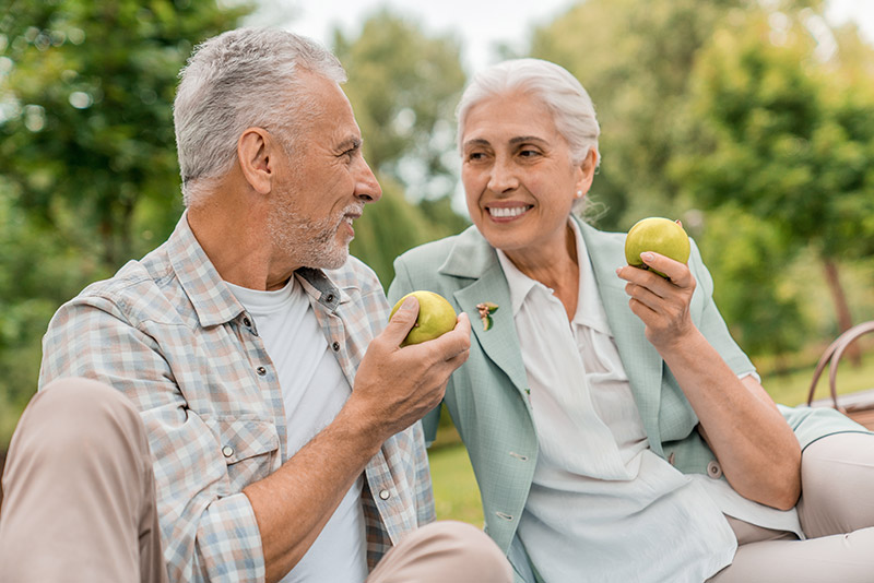 Couple enjoying green apples in a park, smiling and engaging in conversation, reflecting a healthy lifestyle and companionship.